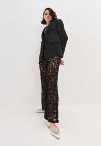 Zwarte blazer met een gladde afwerking, gecombineerd met zwarte kanten broeken met bloemmotieven en zilverkleurige puntige schoenen.