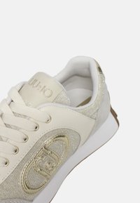 Sneaker con tomaia in pelle color crema, dettagli glitterati argentati e un logo dorato. Presenta lacci bianchi e una suola in gomma testurizzata.