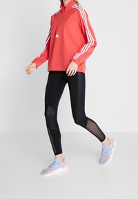 Punainen vetoketjulla varustettu takki, jossa on valkoiset raidat, mustat leggingsit, joissa on verkko-insertit, ja moniväriset lenkkarit, joissa on neulottu yläosa ja kumi-pohja.