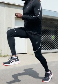 Czarny sportowy top z długim rękawem z białymi akcentami, czarne legginsy i czarne trampki z czerwonymi detalami, prezentujące ruch biegowy na świeżym powietrzu.