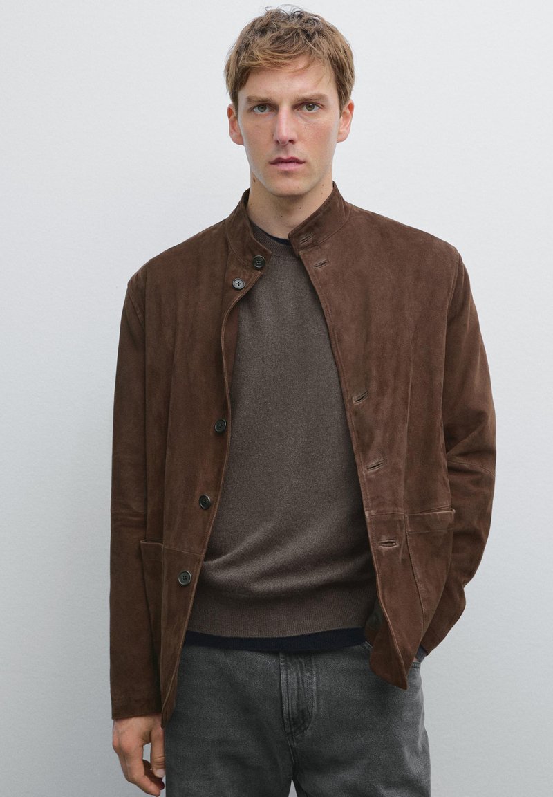 Veste en daim marron avec un col haut, fermeture à boutons et poches. Portée sur un pull marron foncé, associée à un pantalon gris.