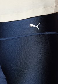 Granatowe legginsy o gładkiej, elastycznej teksturze, z srebnym logo Pumy na lewym biodrze. Zaprojektowane do odzieży sportowej, dopasowany krój.