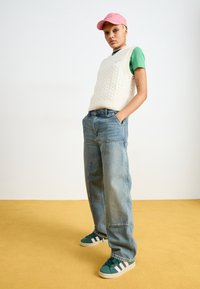 Gilet in maglia bianca a trecce, t-shirt verde, jeans baggy azzurri chiari, sneakers verdi con strisce rosa e un cappellino rosa su uno sfondo neutro.
