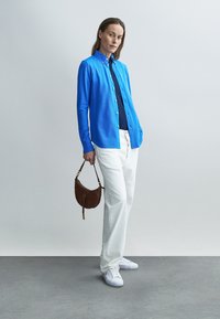 Niebieska koszula na guziki na ciemnej bluzce, białe spodnie i białe trampki. Mała brązowa torebka na pasku dopełnia strój.