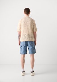 Strikket beige t-skjortegenser med åpen vevdesign, kombinert med blå denim cargo-shorts med lap pockets og dekorativ søm.