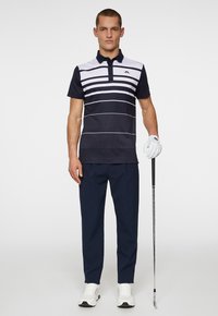 Marinblå och vit randig golfpolo med krage, ihop med marinblå byxor och vita skor. Modellen håller en golfklubba och bär en vit handske.