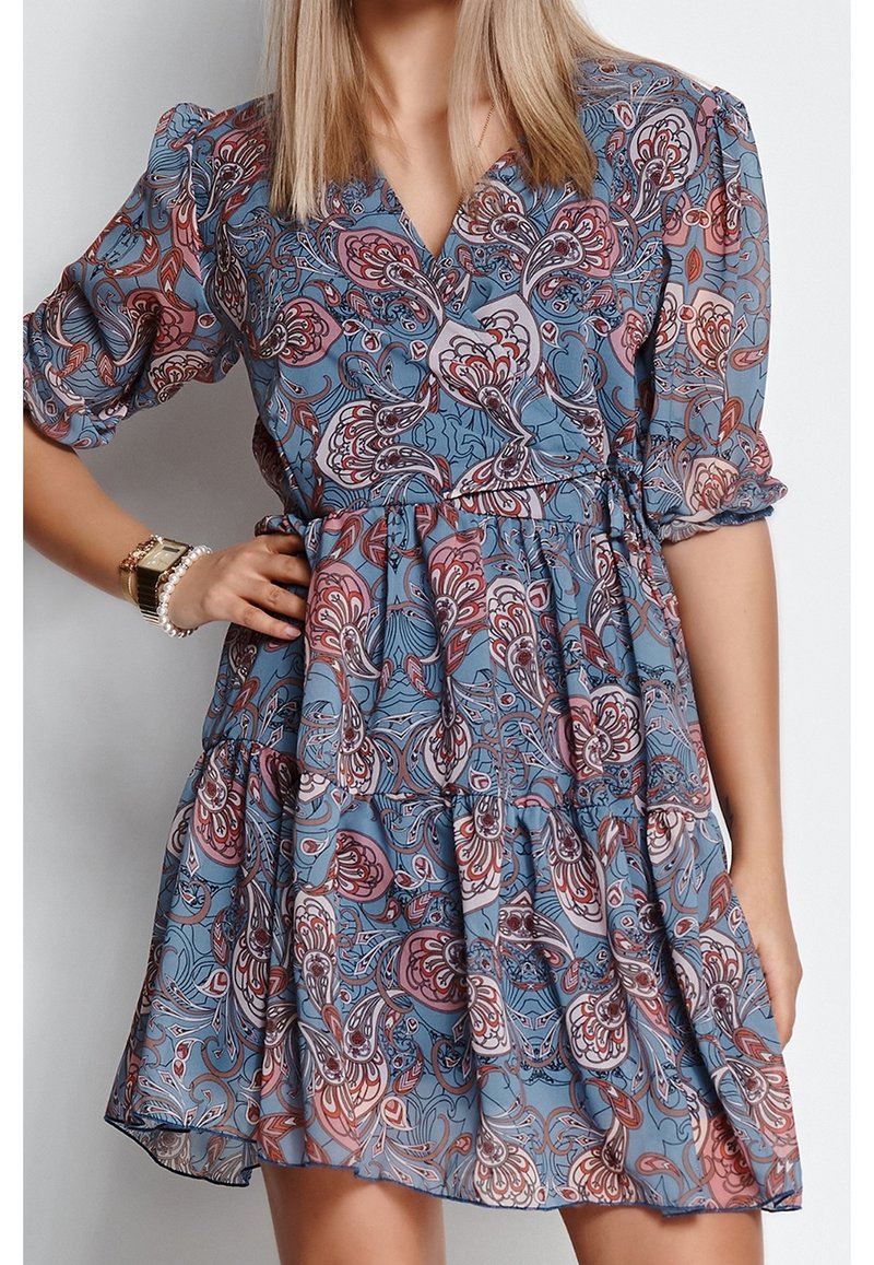 Femme portant une robe bleue avec des motifs floraux roses et rouges, des manches bouffantes et une taille nouée, avec une montre en or et un bracelet de perles.