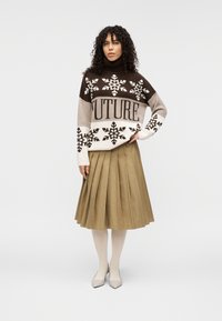 Pull en tricot marron et crème avec des motifs de flocons de neige et le texte « FUTURE », associé à une jupe plissée verte et des chaussures beiges pointues.