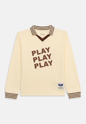 Kremfarvet langærmet skjorte med brunstribet krave og manchetter, med brun "PLAY PLAY PLAY" tekst og en lille grafisk patch nær kanten.