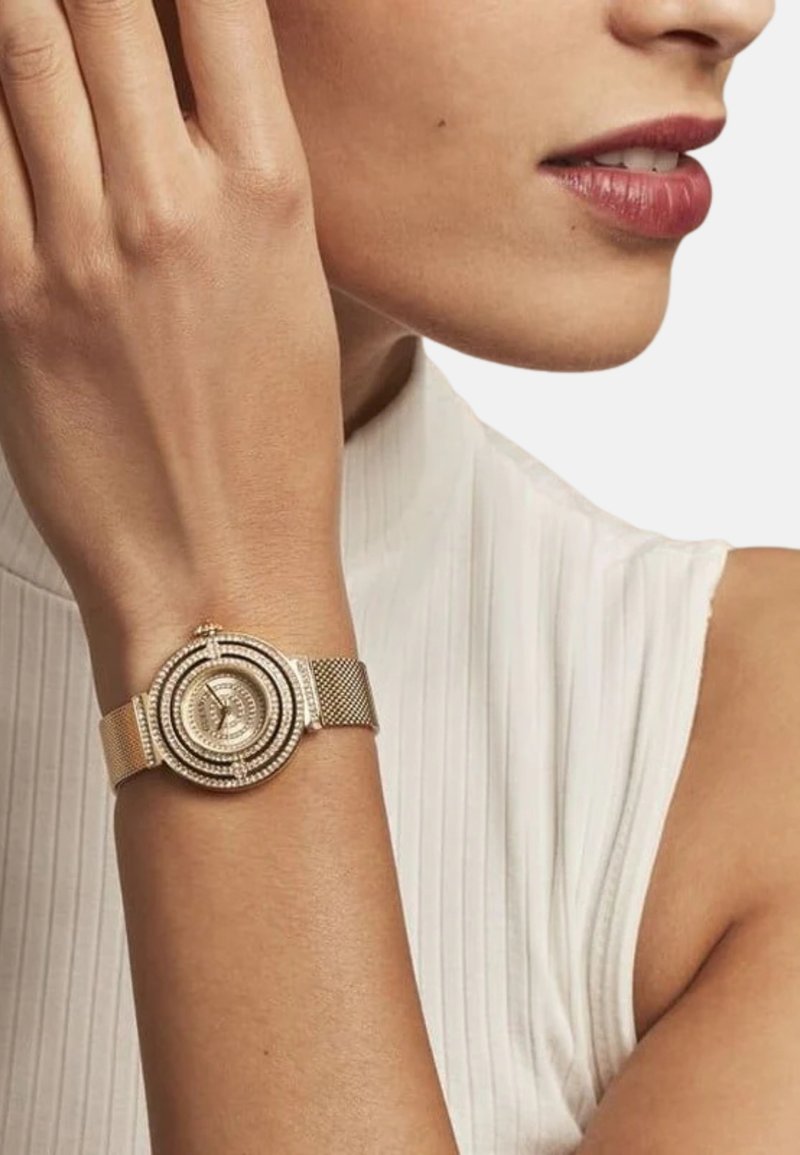 Zlatni mrežasti sat s okruglim licem koji ima koncentrične prstenove i prozirne kamenčiće. Prikazan na golom zapešću na bijeloj pozadini.