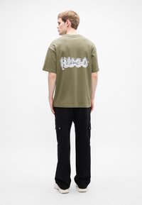 Mees, seljaga, seljas oliiviroheline HUGO logo T-särk, mustad cargo-püksid ja valged tossud, tausta on ühtlane valge.
