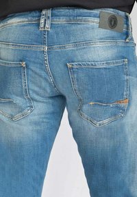 Jean en denim bleu avec une texture légèrement décolorée, doté de deux poches arrière et d'une étiquette de marque grise sur la ceinture.