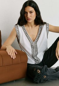 Kvinde med langt mørkt hår iført en hvid ærmeløs top med perledekorationer sidder på gulvet ved siden af en brun polstret stol.