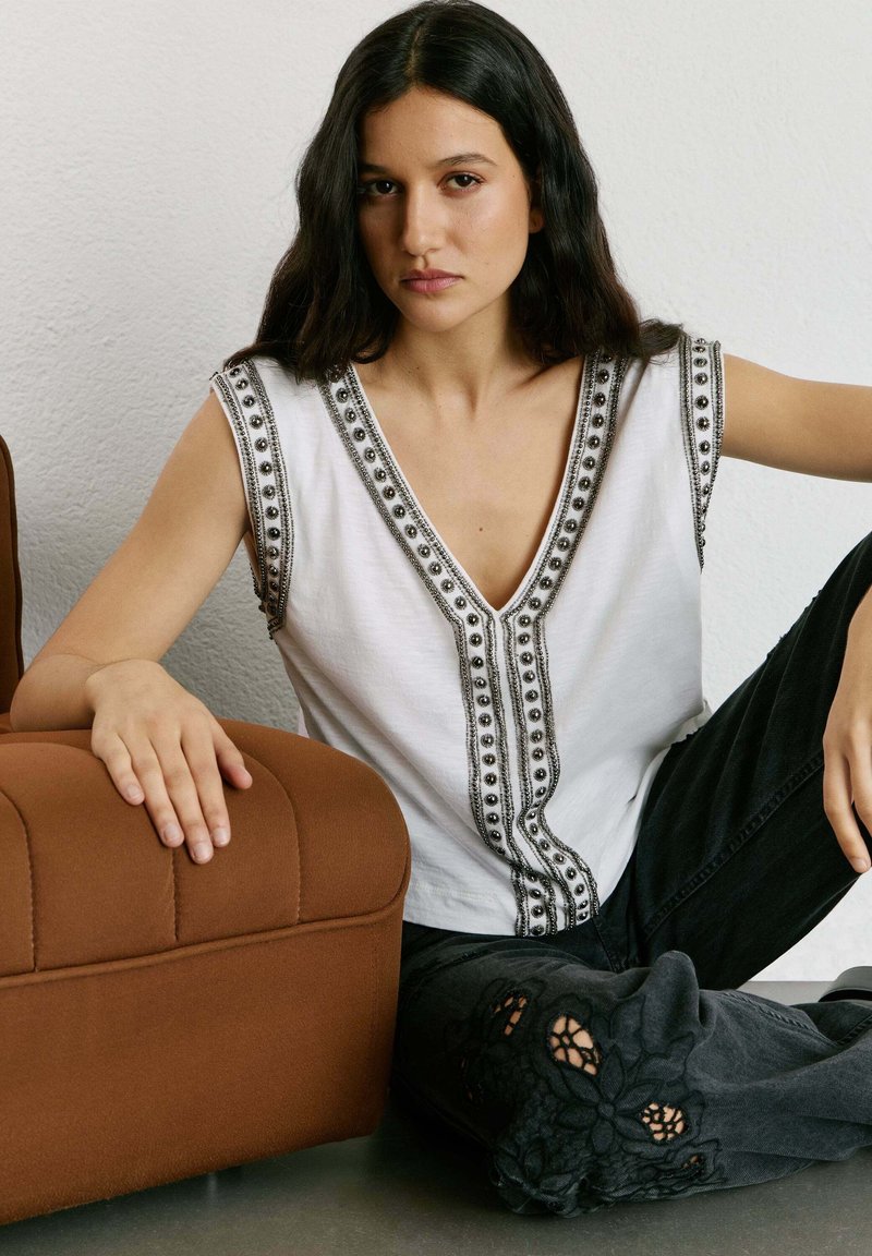 Kobieta z długimi ciemnymi włosami, ubrana w białą, bezrękawnikową bluzkę z koralikowymi zdobieniami, siedzi na podłodze obok brązowego, miękkiego fotela.