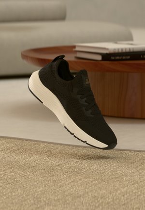 Schwarzer Strick-Sneaker mit weißer Sohle, der über einem beigen Teppich, einem hölzernen Couchtisch und gestapelten Büchern in einem neutral gehaltenen Raum schwebt.