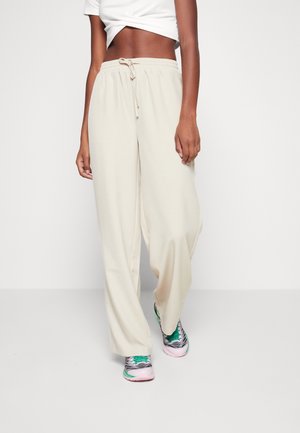 Person iført løse beige bukser med snor, hvid crop-top og pink, grønne og grå sneakers mod en ensfarvet hvid baggrund.