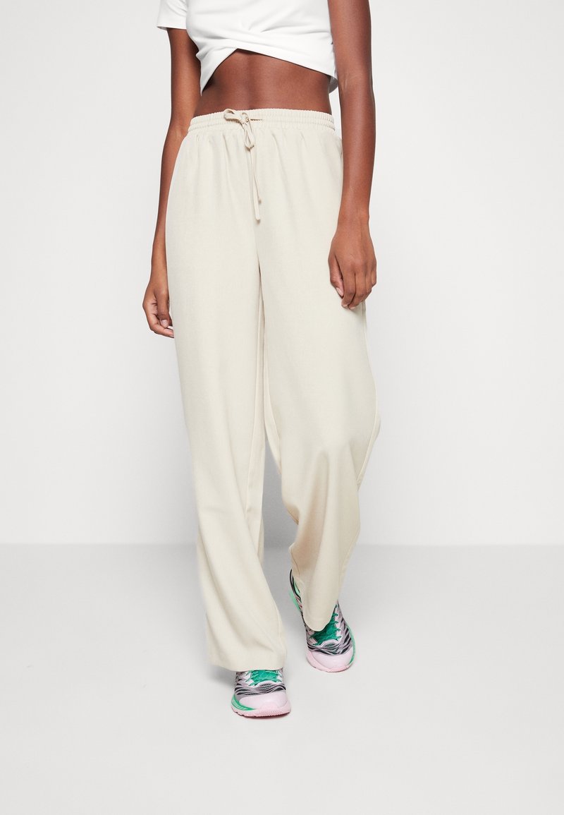 Personne portant un pantalon beige ample avec cordon, un crop top blanc, et des baskets roses, vertes et grises sur un fond blanc uni.