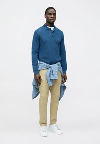 Blå langermet pikéskjorte, khakifargede bukser og hvite sneakers. Lyseblå skjorte knyttet rundt livet. Minimalistisk design, uten mønster.