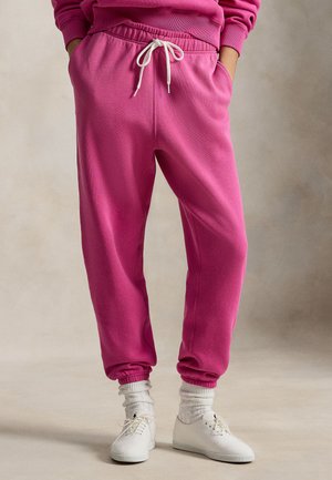 Różowe spodnie dresowe z elastycznym pasem i białym sznurkiem do związania, o luźnym kroju i ściągaczach przy nogawkach. Noszone z białymi trampkami i skarpetkami.