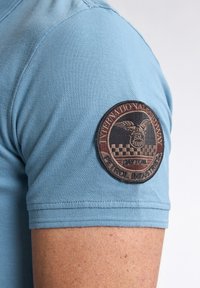 Vaaleansininen lyhytnhihainen paita, jossa on pyöreä, ruskea brodeerattu merkki, jossa teksti "International Speedway, Daytona, Petrol Industries."