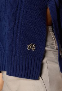 Tengerészkék kötött pulóver kiemelkedő textúrával, bordázott mandzsettákkal és kis fémszínű logóval az alsó szegélyen.
