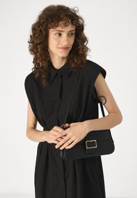 Μαύρη ορθογώνια τσάντα χεριού με λεία υφή και χρυσό κούμπωμα, φορεμένη χιαστί με ένα μαύρο πουκάμισο που διαθέτει δομημένο γιακά.