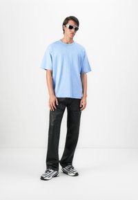 Young man wearing white sunglasses, light blue oversized t-shirt, black cargo pants, and silver-white sneakers, standing against a white background.