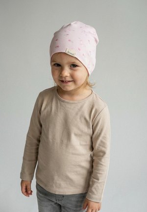 Young child wearing a light pink beanie with small floral prints, beige long-sleeve shirt, and gray pants against a plain background.