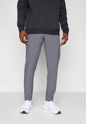 Homme portant un pantalon slim gris, un pull noir et des baskets blanches debout devant un fond uni.