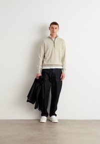 Um suéter bege claro com fecho zip, calças de ganga escuras e ténis brancos; segurando uma jaqueta preta; design minimalista com uma textura suave.