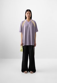 Il modello è in piedi contro uno sfondo semplice, indossando una maglietta sportiva larga di colore lavanda, pantaloni neri larghi e tiene in mano una piccola borsa gialla.