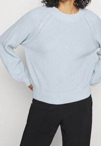 Ljusblå stickad tröja med ribbat fåll och raglanärmar; har öppna sömmar nära axlarna och strukturerad tyg.