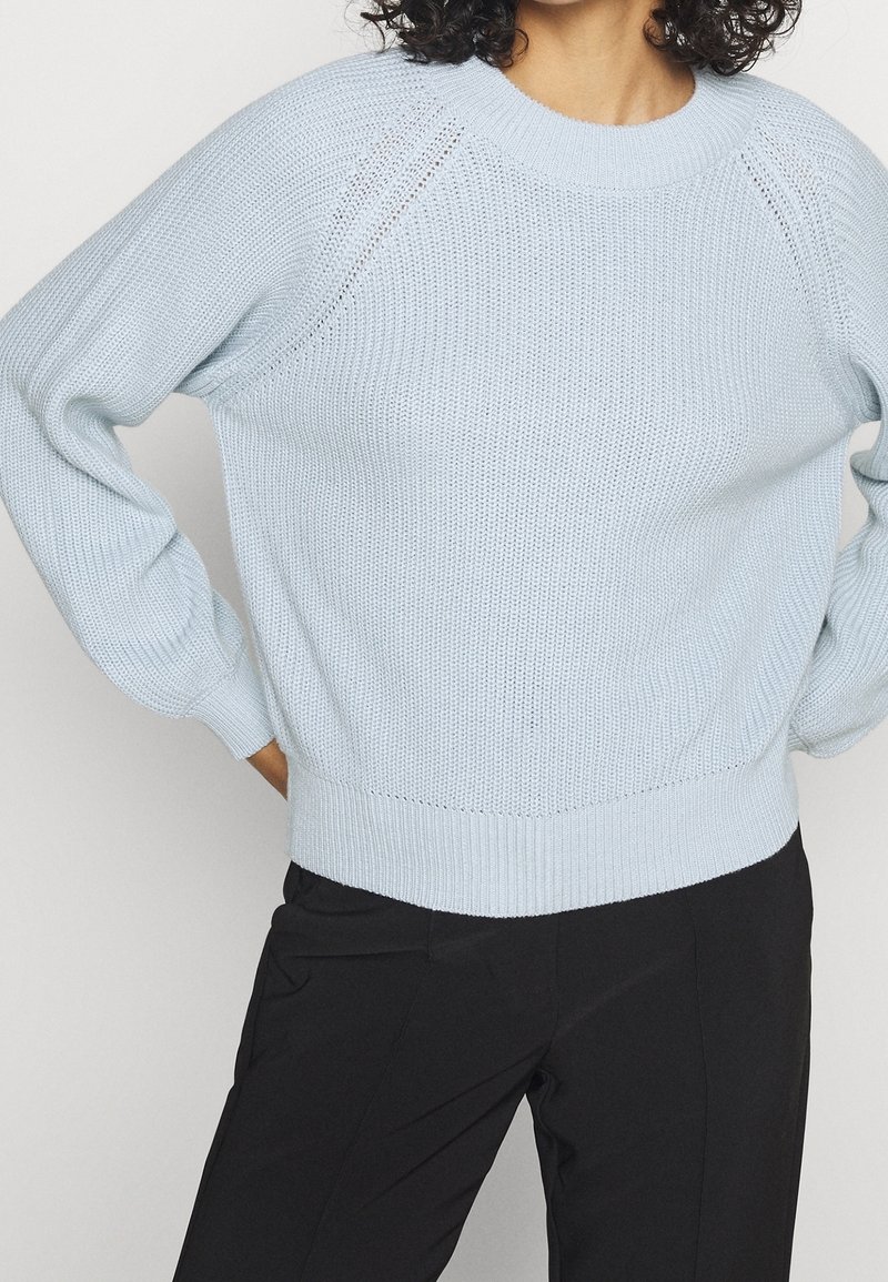 Ljusblå stickad tröja med ribbat fåll och raglanärmar; har öppna sömmar nära axlarna och strukturerad tyg.