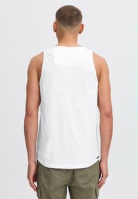Hvid bomulds tanktop med en rund kant, der viser en ensfarvet ryg med en diskret lapdetalje, parret med olivengrønne cargo shorts.