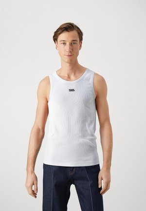 Top de tirantes blanco con textura acanalada y escote redondo, que presenta un pequeño logo negro en el centro. Combinado con unos vaqueros azul oscuro.