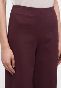 Gros plan d'une personne portant un pantalon large taille haute bordeaux foncé et un haut en maille beige clair, les mains détendues sur les côtés.