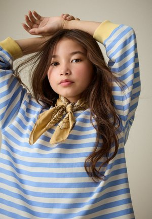 Zils un balts svītrainais pārmēru lielais džemperis ar dzeltenām aprocēm, kopā ar brūnu rakstainu šalli, kas saistīta ap kaklu. Garas viļņotas matu cirtas.