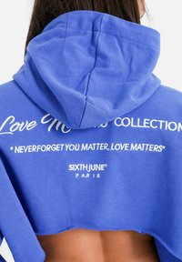 Ingekorte blauwe hoodie met een textuurafwerking, voorzien van witte geborduurde tekst op de achterkant. Logo "SIXTHJUNE PARIS" weergegeven nabij de zoom.