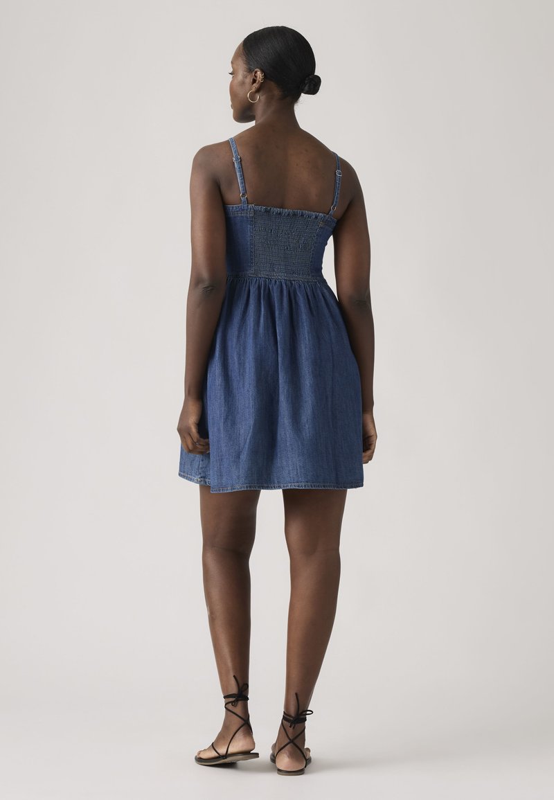 Woman with dark skin wearing a blue sleeveless denim dress and black lace-up flat sandals, standing facing away on a plain background.