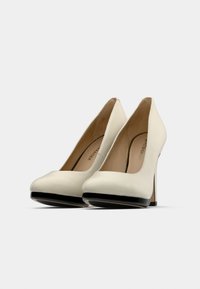 Λευκές ψηλές γόβες με λείο, ματ φινίρισμα. Στρογγυλεμένη σχεδίαση μύτης και  μαύρη λεπτομέρεια στη σόλα. Λεπτό τακούνι στιλέτο και ορατή εσωτερική επένδυση.