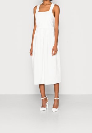 Kobieta ubrana w białą, bezrękawnikową sukienkę midi z kwadratowym dekoltem oraz białe szpilki z paskiem wokół kostki, stojąca na jasnym tle.