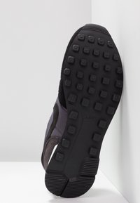Semelle d'une basket noire Nike montrant une semelle extérieure en caoutchouc texturée avec des crampons carrés et des motifs de bande de roulement sur fond blanc.