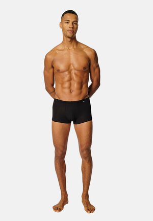 Durchtrainierter Mann steht barfuß vor einfarbigem Hintergrund, trägt schwarze Boxershorts, hat die Hände hinter dem Rücken und einen neutralen Gesichtsausdruck.