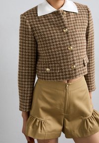 Bruin houndstooth cropped jack met gouden knopen en witte kraag, gedragen met hoge beige shorts met een rits aan de voorkant en ruches.