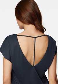 Blusa blu navy con scollatura a V profonda sul retro e maniche corte. Presenta una sottile tracolla che collega le spalle. Tessuto liscio con una leggera texture.