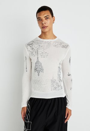 Jeune personne portant une chemise blanche à manches longues avec des croquis architecturaux noirs et du texte, associée à un pantalon noir avec des motifs géométriques blancs.