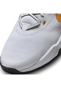 Zapatilla deportiva blanca con parte superior de malla texturizada, acentos grises, logo naranja y suela de goma negra. Diseño con cordones y puntera reforzada.