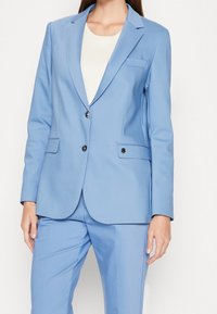 Světlomodrý oblekový sako s výstřihem, dvěma černými knoflíky a bočními kapsami. Vyrobeno z hladkého materiálu s strukturovaným střihem.