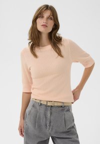 Eine Frau mit hellbraunen Haaren trägt ein blasspfirsichfarbenes Strickoberteil mit Ellenbogenärmeln und einen hellbeigen Gürtel über hochgeschnittenen grauen Jeans, während sie mit einer Hand auf der Hüfte posiert.