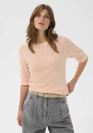 Eine Frau mit hellbraunen Haaren trägt ein blasspfirsichfarbenes Strickoberteil mit Ellenbogenärmeln und einen hellbeigen Gürtel über hochgeschnittenen grauen Jeans, während sie mit einer Hand auf der Hüfte posiert.